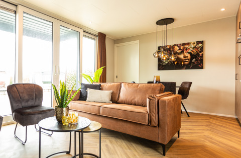 Moderne leefruimte met grote ramen, lederen zetel en muurkunst in Waterhoeve Wellness, Park Westerkogge.