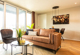 Moderne leefruimte met grote ramen, lederen zetel en muurkunst in Waterhoeve Wellness, Park Westerkogge.