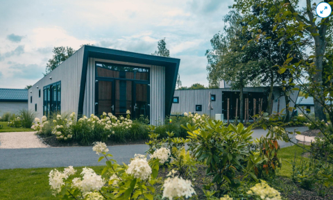 Modern lodge called Just Nature, surrounded by green plants and flowers in a peaceful, natural setting.