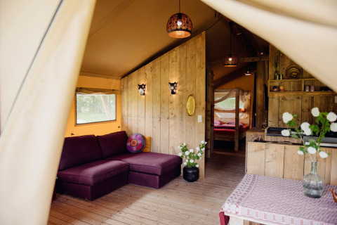 Interno di una tenda safari di lusso con pannelli in legno, divano viola, fiori e illuminazione accogliente.