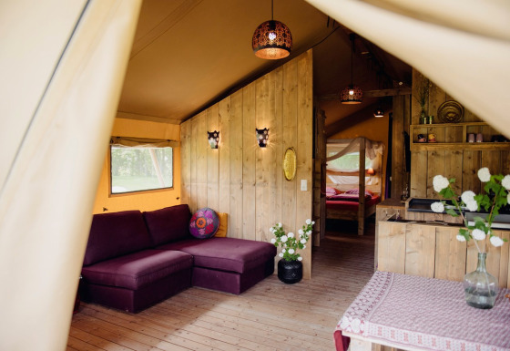 Interno di una tenda safari di lusso con pannelli in legno, divano viola, fiori e illuminazione accogliente.