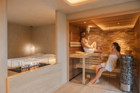 Suite de bienestar en Woodz Lodges, Bélgica, con dos personas en sauna y una moderna chimenea interior.