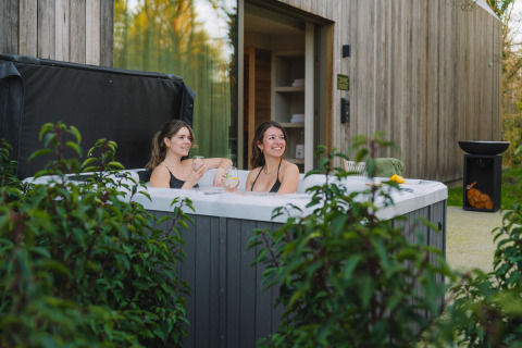 Two women relax in an outdoor hot tub at the Wellness Suite, Woodz Lodges, Belgium, surrounded by nature.
