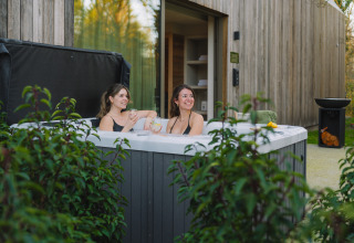 Dos mujeres se relajan en un jacuzzi exterior en Wellness Suite, Woodz Lodges, Bélgica, rodeadas de naturaleza.