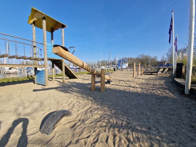 Parque infantil con balancín, tobogán y puente de cuerda en arena junto a un glamping en día soleado.