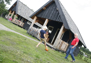 Niño y adulto juegan fútbol frente a cabañas negras en Wood Lodge, Holiday park De Boshoek, Países Bajos.