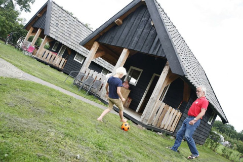 Child and adult play soccer on grass in front of black wooden cabins at Wood Lodge, Holiday park De Boshoek, Netherlands.