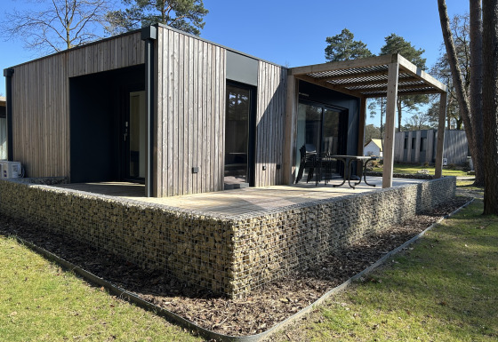 Domus 4+2 lodge met hedendaagse houten buitenzijde, ruim terras en buitenzitje in een groene omgeving.