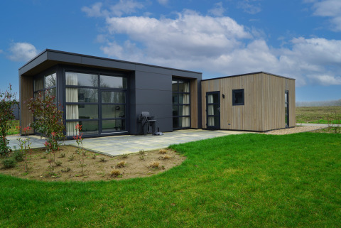 Moderne glampingaccommodatie in een landelijk veld met grote ramen, groen gazon en blauwe lucht.