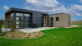 Moderne glampingverblijf op het platteland met grote ramen, terras en groene grasmat onder blauwe lucht.