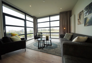 Moderne glamping woonkamer met grote ramen, comfortabele grijze banken en uitzicht op het landschap.