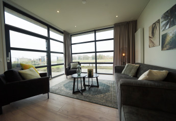 Moderne glampingstue med store vinduer, grå sofaer, grønne puder og naturskøn udsigt udenfor.