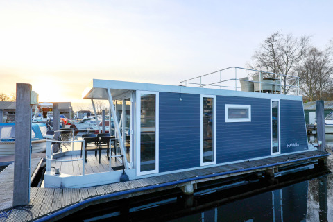 Husbåd ved marina med moderne design, blå facade, terrasse og tagterrasse, set i Holland ved solnedgang.