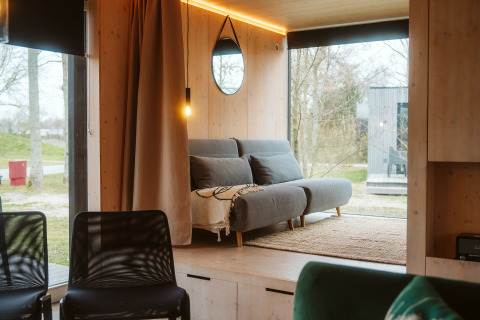 Modern houten hutinterieur met grijze bank, grote ramen en uitzicht op de natuur in een Nederlands park.