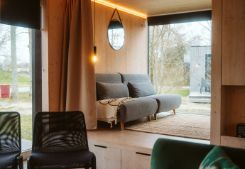 Moderne træhytte interiør med gråt sofaområde, store vinduer og naturskøn udsigt i Nederlandene.