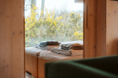 Bed bij raam met uitzicht op bloeiende struiken, in een houten Ark Shelter op De Klepperstee, Nederland.