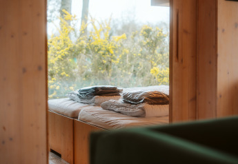 Bed bij raam met uitzicht op bloeiende struiken, in een houten Ark Shelter op De Klepperstee, Nederland.