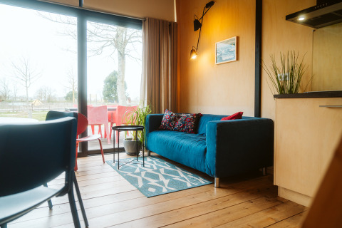 Woonkamer met blauwe bank, kleurrijke kussens en groot raam in The Hoagte, Holiday park De Klepperstee.