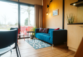 Wohnzimmer mit blauem Sofa, bunten Kissen und großem Fenster im Tiny House im Holiday Park De Klepperstee.