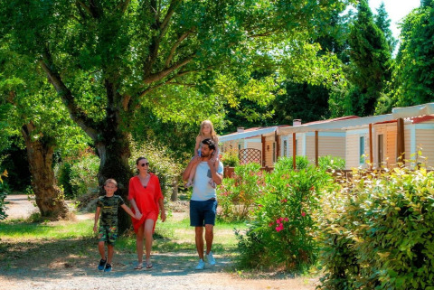 Una famiglia cammina insieme davanti a casette immerse nel verde tra alberi e cespugli in una giornata di sole.
