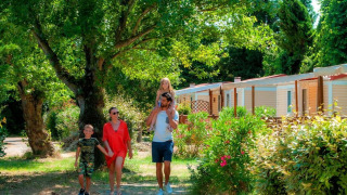 Una familia pasea frente a acogedoras cabañas rodeadas de árboles y arbustos verdes en un día soleado.
