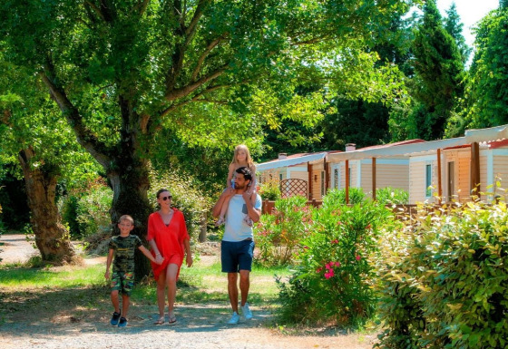 Una famiglia cammina insieme davanti a casette immerse nel verde tra alberi e cespugli in una giornata di sole.