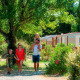 Una famiglia cammina insieme davanti a casette immerse nel verde tra alberi e cespugli in una giornata di sole.