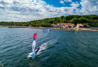 Windsurfer gleiten über das Wasser nahe grüner Hügel und Gebäude bei Sigean, Occitanie, Frankreich.