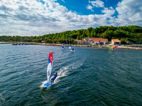 Windsurfer gleiten über das Wasser nahe grüner Hügel und Gebäude bei Sigean, Occitanie, Frankreich.