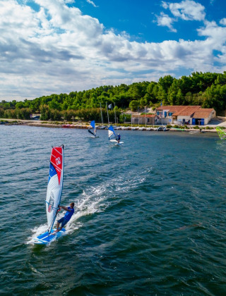 Windsurfers glide on the sea with coastal houses and lush green hills near Sigean, Occitanie, France.