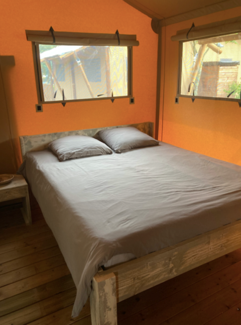 Cama dentro de la tienda safari Wood lodge en Camping Seasonova Ensoya, Francia, con marco de madera y ventanas.