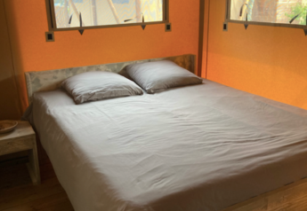 Cama dentro de la tienda safari Wood lodge en Camping Seasonova Ensoya, Francia, con marco de madera y ventanas.