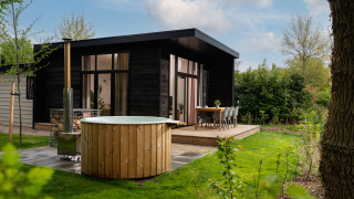 En moderne lodge ved navn Wellness Boutique med udendørs badekar, terrasse og grøn have under blå himmel.
