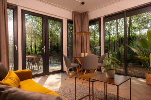 Stilfuld stue i Happy Oak Lodge med store vinduer, moderne møbler og udsigt til naturen udenfor.