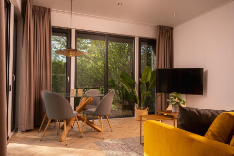Salón moderno de lodge con grandes ventanales, mesa de cristal, sillas grises y sofá amarillo, vista verde.