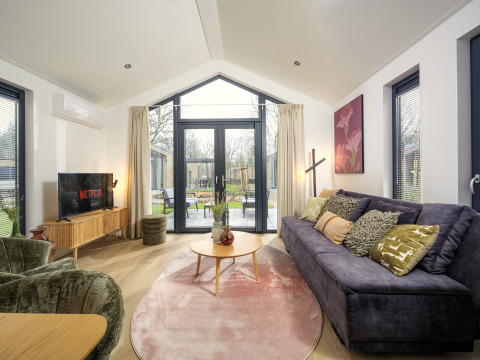 Sala moderna en Tiny Lodge con sofá grande, TV y puertas de vidrio abiertas a la terraza en Veluwepark de Bosgraaf.
