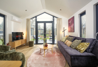 Sala moderna en Tiny Lodge con sofá grande, TV y puertas de vidrio abiertas a la terraza en Veluwepark de Bosgraaf.