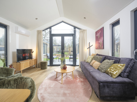Sala moderna en Tiny Lodge con sofá grande, TV y puertas de vidrio abiertas a la terraza en Veluwepark de Bosgraaf.