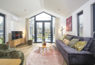 Sala moderna en Tiny Lodge con sofá grande, TV y puertas de vidrio abiertas a la terraza en Veluwepark de Bosgraaf.