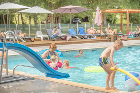 Familier nyder swimmingpoolen i Club Les Ormes feriepark med rutsjebane og solsenge i baggrunden.
