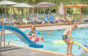 Familier nyder swimmingpoolen i Club Les Ormes feriepark med rutsjebane og solsenge i baggrunden.