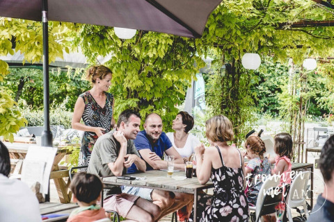 Gruppe voksne og børn nyder sommermåltid under grønne planter på udendørs café i Club Les Ormes, Frankrig.