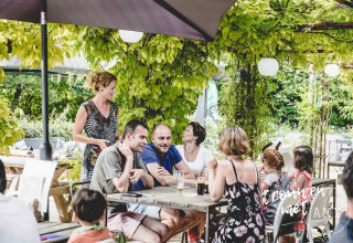 Gruppe voksne og børn nyder sommermåltid under grønne planter på udendørs café i Club Les Ormes, Frankrig.