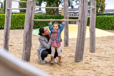 Une mère aide son jeune enfant à jouer sur les barres dans le sable à Berg Lodge - Sauna, Resort Mooi Bemelen.