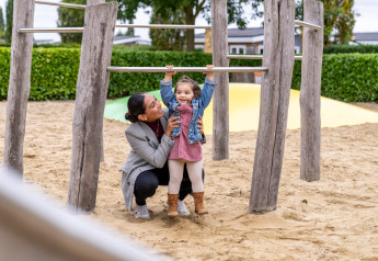 A mother helps her young child play on climbing bars in the sand at Berg Lodge - Sauna, Resort Mooi Bemelen.