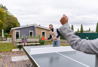 Zwei Männer spielen Tischtennis draußen bei Berg Lodge - Sauna im Resort Mooi Bemelen, Niederlande, mit einem Kind auf der Bank.