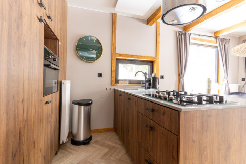 Cocina moderna de madera con estufa de gas, reloj de pared y grandes ventanas en Panorama Lodge, Pressegger See, Austria.