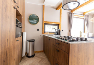 Cocina moderna de madera con estufa de gas, reloj de pared y grandes ventanas en Panorama Lodge, Pressegger See, Austria.