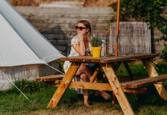 Kvinde sidder ved et træbord under en parasol foran Bell Tent 500 hos REBL Outdoor i Holland.