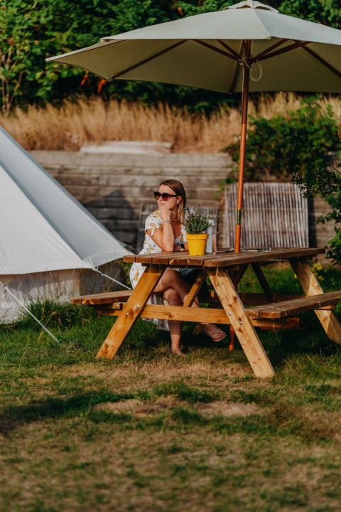 Frau sitzt an einem Holztisch unter einem Sonnenschirm vor dem Bell Tent 500 bei REBL Outdoor, Niederlande.
